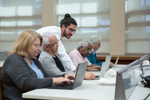 Homem ensinando um grupo de idosos a usar laptops em uma sala de aula.
