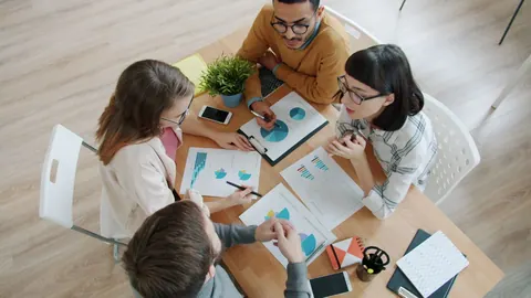 Pessoas diversas trabalhando juntas em uma mesa, analisando gráficos e documentos.
