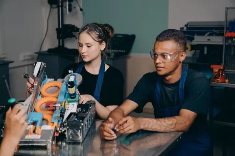 Jovens aprendizes em uma fábrica, focados em uma máquina complexa.