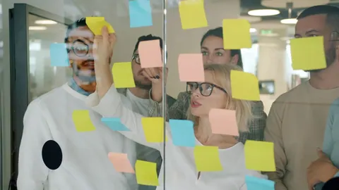 Pessoas trabalhando em equipe, usando post-its em um painel de vidro.