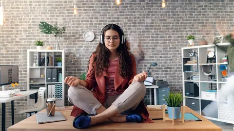 Mulher meditando com fones de ouvido em cima de uma mesa de escritório.
