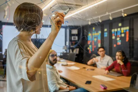 Mulher escrevendo em painel de vidro durante reunião de equipe em escritório moderno.