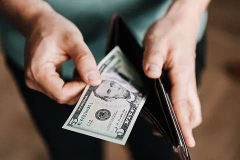 Mãos segurando uma nota de cinco dólares e uma carteira aberta.