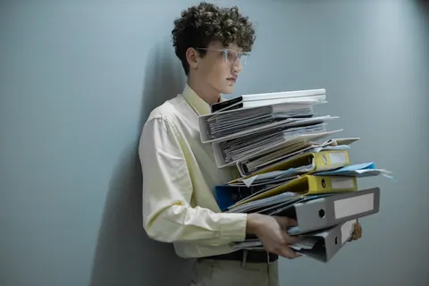 Homem jovem sobrecarregado carregando uma pilha de documentos e pastas.
