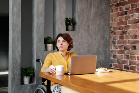 Mulher de camisa amarela sentada em cadeira de rodas, olhando para o lado, com laptop e xícara na mesa.