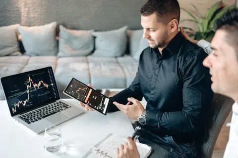Homens analisando gráficos de criptomoedas em tablet e laptop, discutindo dados financeiros.