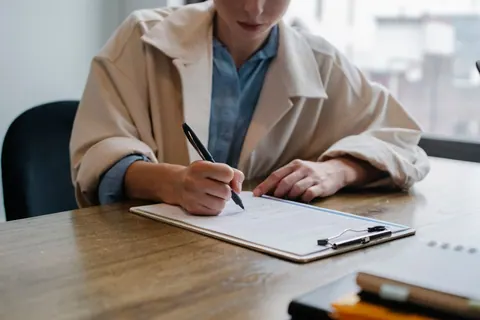 Pessoa escrevendo em uma prancheta com caneta preta sobre uma mesa de madeira.