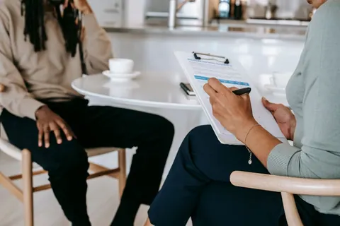 Duas pessoas sentadas em uma mesa branca, uma escrevendo em uma prancheta.