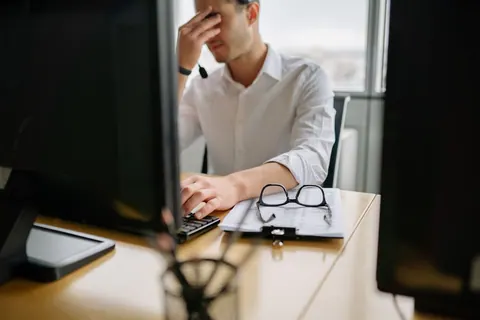 Homem no escritório com a mão no rosto, parecendo exausto ou com dor de cabeça.