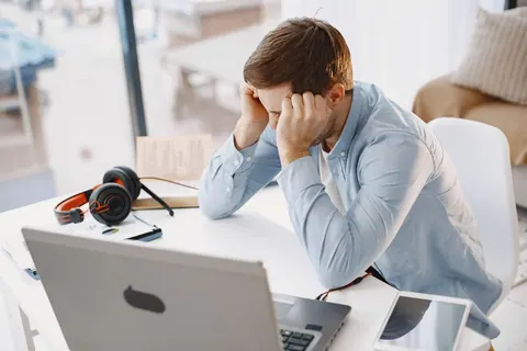 Homem frustrado no escritório em casa, com as mãos na cabeça, olhando para o laptop.