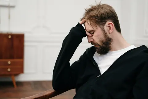 Homem com blazer preto sentado em cadeira, com a mão na testa, pensativo.