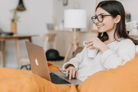 Mulher sorridente usando laptop e tomando café, em ambiente doméstico e confortável.