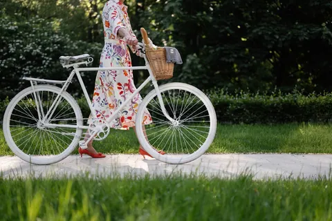 Pessoa em vestido floral e sapatos vermelhos empurrando uma bicicleta branca com cesta.