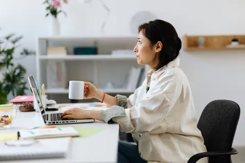 Mulher asiática trabalhando em laptop, segurando uma xícara, com expressão pensativa.
