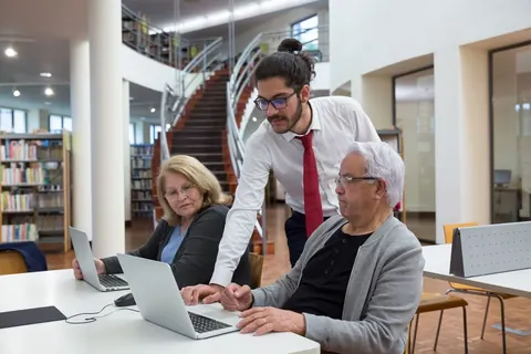 Homem ensinando idosos a usar um laptop em uma sala de aula.