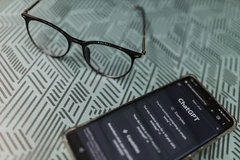 Óculos de grau escuros e um celular com o aplicativo ChatGPT aberto sobre uma superfície estampada.