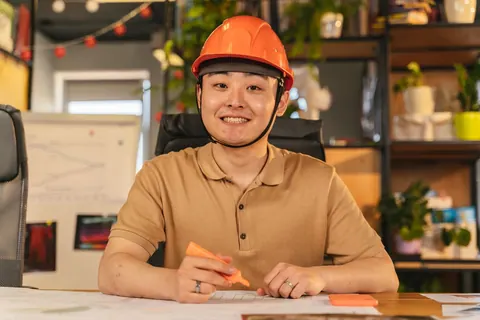 Homem asiático sorrindo, de capacete, sentado à mesa com caneta marca-texto.
