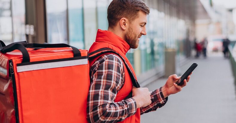 Homem branco com mochila de entregador de aplicativos segurando um celular e sorrindo